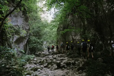 Un gruppo di escursionisti con caschi gialli attraversa un suggestivo canyon in Italia, tra rocce e vegetazione lussureggiante.