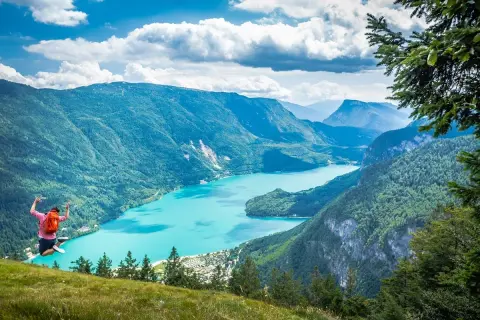 Giovane salta felice con zaino rosso, godendosi le passeggiate facili vicino a Trento con vista su lago turchese e montagne verdi.