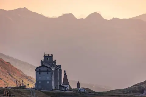 Edificio imponente tra paesaggi di montagna bellissimi, con cime sfumate all'orizzonte e un cielo rosato.