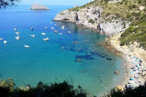 La cala del Gesso, Monte Argentario, offre un mare cristallino con barche all'ancora e una spiaggia affollata di bagnanti sotto ombrelloni colorati.