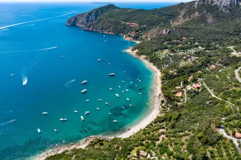 Vista aerea di una baia con spiagge dorate e acque cristalline, un vero gioiello della cartina della Toscana. Diverse imbarcazioni solcano il mare blu intenso.