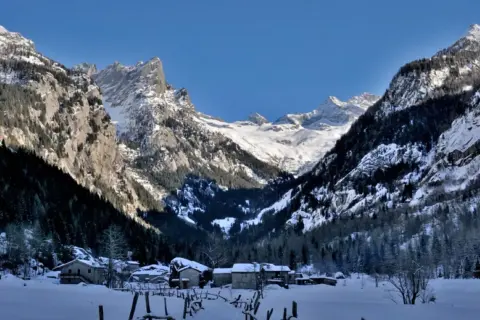 Case inneggiate nel Val di Mello inverno, con montagne imponenti e case di pietra sotto un cielo azzurro.