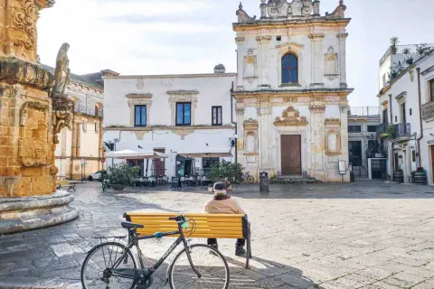 Un uomo seduto su una panchina in una piazza, con una bicicletta accanto. Architettura barocca e scorci dei borghi più belli del Salento.