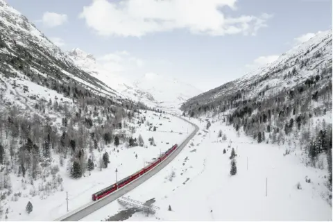 Un treno rosso attraversa una valle innevata, un viaggio tra i **treni panoramici Italia** con montagne maestose e alberi spogli.