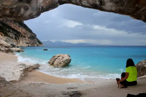 Vista dalla grotta sulla spiaggia di Cala Goloritzè, raggiungibile con un **percorso a piedi**. Una donna con un cane ammira il mare turchese.