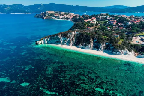 Arcipelago Toscano: una costa con scogliere bianche, spiaggia dorata e mare cristallino, con un borgo storico in lontananza. Un gioiello tra le isole visitabili.