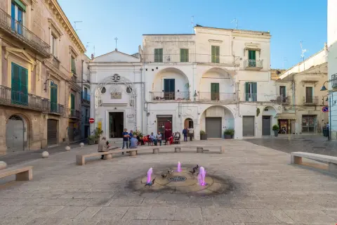 Piazza nel gravina in puglia centro storico con fontana rosa e persone sedute.