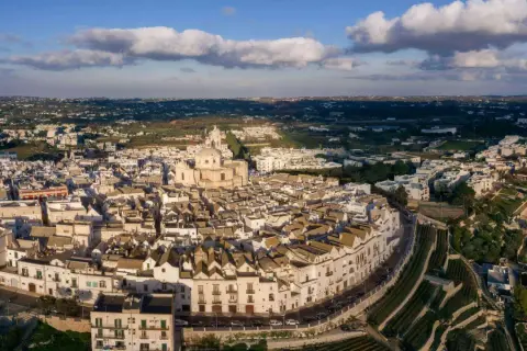 Vista aerea di Locorotondo di sera, con le sue case bianche a tetto spiovente e la chiesa principale che domina il paesaggio collinare.
