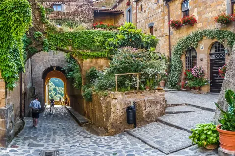 Un vicolo acciottolato in un borgo da visitare, con case in pietra ricoperte di edera e fiori. Un arco conduce a una vista panoramica.