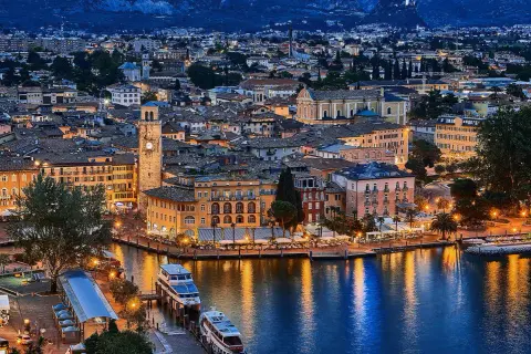 Incantevole vista serale di un borgo sul lago, con edifici colorati, un campanile e barche illuminate. Un gioiello tra i paesi del Trentino Alto Adige.