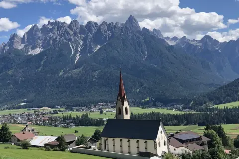 Chiesa con campanile rosso in primo piano, sullo sfondo le maestose vette delle Dolomiti e i **paesi più belli** della **Val Pusteria**.