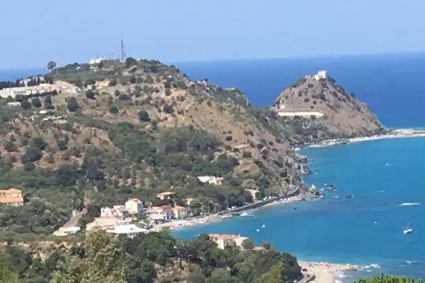 Panoramica di Barcellona Pozzo di Gotto con case bianche e rosse affacciate sul mare blu intenso, colline verdi e un castello in cima.