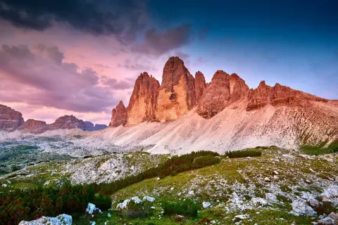 Le maestose cime di Lavaredo illuminate dal tramonto, con un cielo viola e blu che fa da sfondo a un paesaggio alpino mozzafiato.