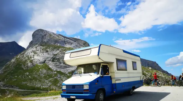 Camper blu e bianco parcheggiato in montagna, pronto per esplorare gli **itinerari camper Italia**. Due ciclisti sullo sfondo.