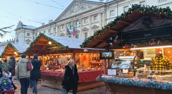 Mercatini di Natale Aosta: bancarelle addobbate con luci e ghirlande, prodotti tipici e gente che passeggia. Atmosfera magica.
