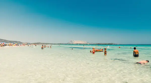 Spiaggia affollata in Sardegna, tra le zone più belle, con gente che nuota e si rilassa nell'acqua cristallina.