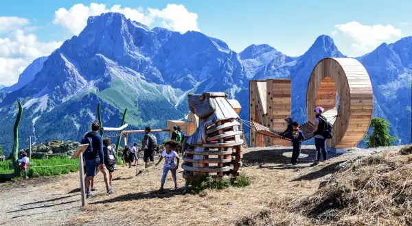 Un'estate a San Martino di Castrozza: bambini giocano in un parco avventura con sculture in legno e montagne maestose sullo sfondo.