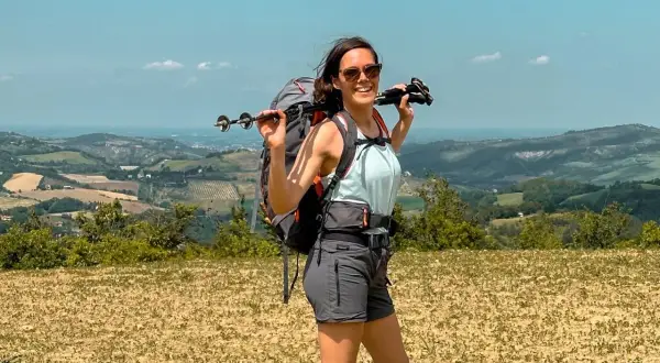 Donna con zaino e bastoncini da trekking, sorride con vista panoramica. Un'escursionista pronta per le tappe del Cammino degli Dei.