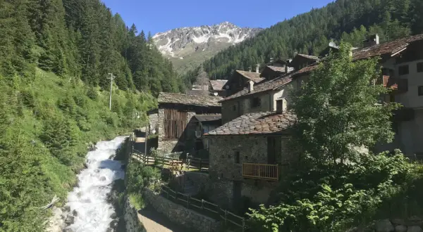 Un pittoresco borgo alpino nel Nord Italia, con case in pietra e tetti in ardesia, incastonato tra boschi rigogliosi e un torrente impetuoso.