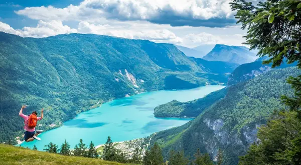 Giovane salta felice con zaino rosso, godendosi le passeggiate facili vicino a Trento con vista su lago turchese e montagne verdi.
