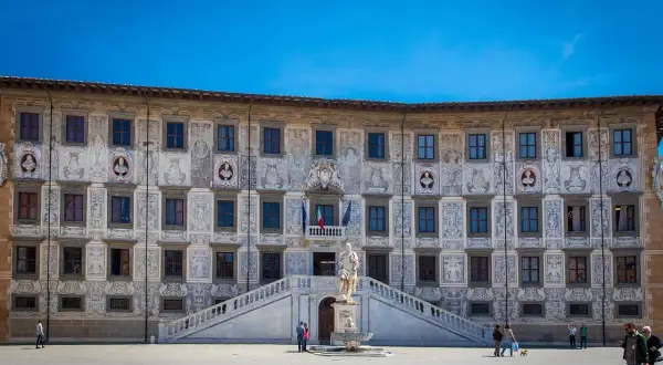 Palazzo della Sapienza a Pisa, con la sua facciata decorata, si affaccia su una piazza dove si intravede la **torre di Pisa** in lontananza, nella **Piazza del Duomo, Pisa, PI**.