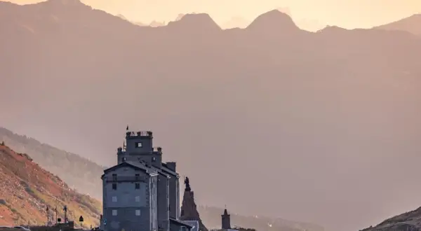 Edificio imponente tra paesaggi di montagna bellissimi, con cime sfumate all'orizzonte e un cielo rosato.