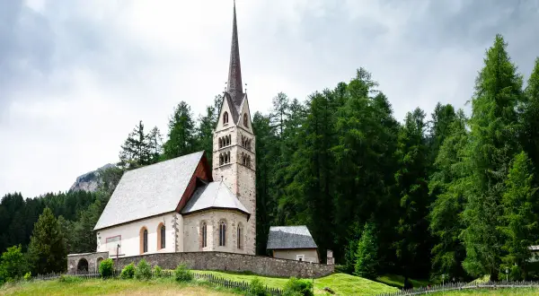 Chiesa di San Giovanni di Fassa, con campanile a cuspide, circondata da un fitto bosco di abeti e montagne sullo sfondo.