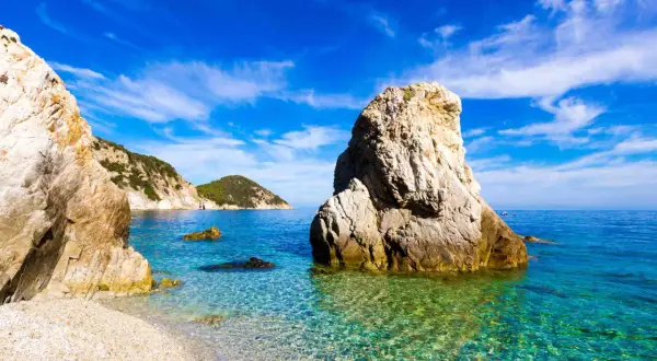 Scogli imponenti e un mare cristallino toscana, con acque turchesi che lambiscono una spiaggia di ciottoli bianchi sotto un cielo azzurro.