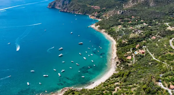 Vista aerea di una baia con spiagge dorate e acque cristalline, un vero gioiello della cartina della Toscana. Diverse imbarcazioni solcano il mare blu intenso.