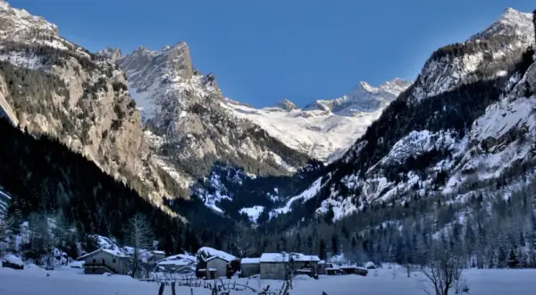 Case inneggiate nel Val di Mello inverno, con montagne imponenti e case di pietra sotto un cielo azzurro.