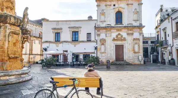 Un uomo seduto su una panchina in una piazza, con una bicicletta accanto. Architettura barocca e scorci dei borghi più belli del Salento.