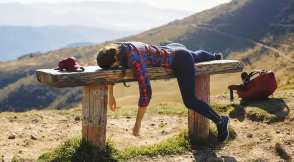 Donna esausta si riposa su una panca in montagna dopo un'intensa attività. Zaino e cappellino rossi accanto a lei.