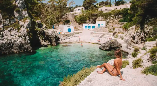 Una donna ammira le acque cristalline di una baia pugliese, con piccole spiagge sabbiose e case bianche arroccate sulla scogliera.