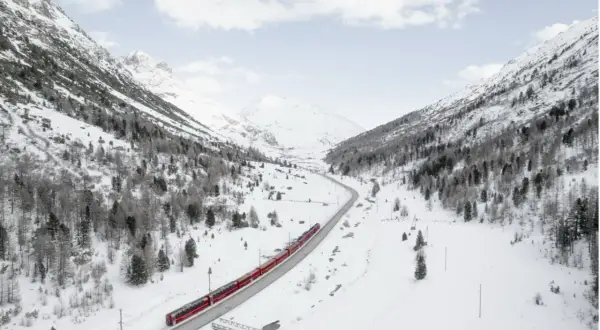 Un treno rosso attraversa una valle innevata, un viaggio tra i **treni panoramici Italia** con montagne maestose e alberi spogli.