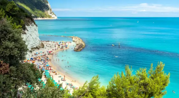 Spiaggia affollata con ombrelloni colorati e gente che prende il sole, bagnata da un **abruzzo mare bello** e cristallino, con scogliere bianche e vegetazione rigogliosa.