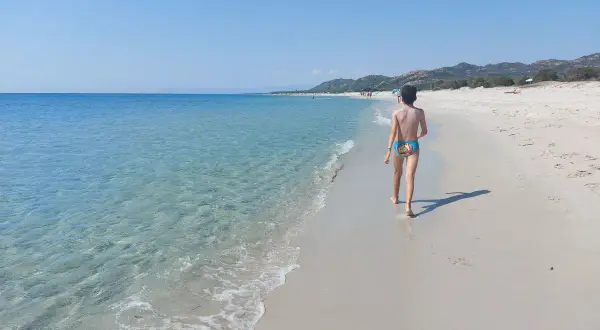 Bambino cammina sulla spiaggia della costa est Sardegna, mare cristallino e cielo azzurro.
