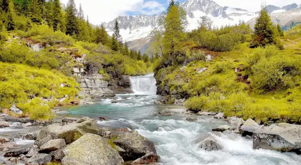 Cascata alpina e torrente cristallino, perfetti per gite in Trentino in giornata. Montagne innevate sullo sfondo.