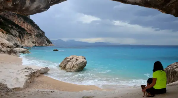 Vista dalla grotta sulla spiaggia di Cala Goloritzè, raggiungibile con un **percorso a piedi**. Una donna con un cane ammira il mare turchese.