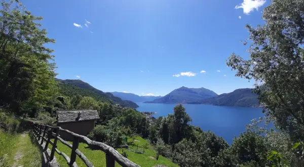 Il sentiero del viandante si snoda tra alberi verdi, con una vista mozzafiato su un lago azzurro e montagne sullo sfondo. Una piccola casa con tetto in pietra si intravede.
