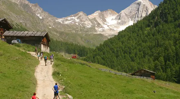Escursionisti e ciclisti percorrono un sentiero nel **Val di Fosse**, tra baite in legno e montagne innevate.