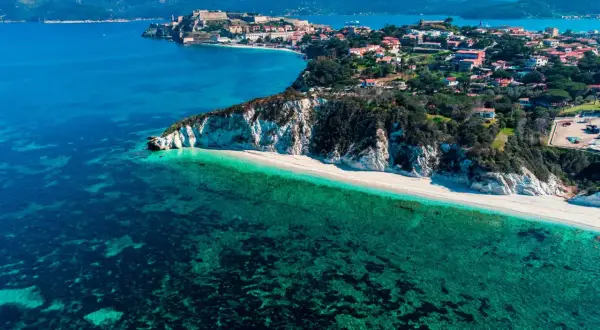 Arcipelago Toscano: una costa con scogliere bianche, spiaggia dorata e mare cristallino, con un borgo storico in lontananza. Un gioiello tra le isole visitabili.