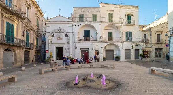 Piazza nel gravina in puglia centro storico con fontana rosa e persone sedute.