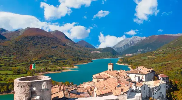 Borgo medievale con torre e lago turchese, un incantevole scorcio di cosa vedere in Abruzzo, tra montagne autunnali e cielo azzurro.
