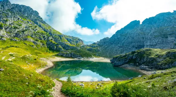 Sentiero in montagna tra verdi prati e rocce, con un lago alpino che riflette il cielo azzurro e nuvole bianche. Splendidi paesaggi naturali.