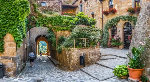 Un vicolo acciottolato in un borgo da visitare, con case in pietra ricoperte di edera e fiori. Un arco conduce a una vista panoramica.