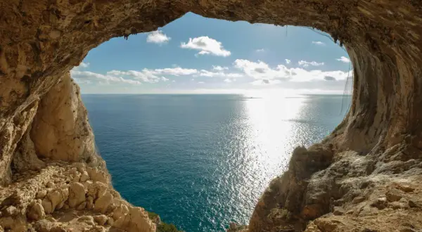 Vista dal mare da una grotta, con un sentiero del pellegrino che si intravede tra le rocce. Il sole brilla sull'acqua.