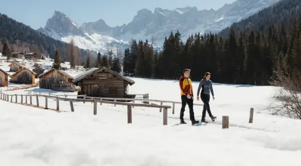 Coppia in cammino tra baite innevate e montagne maestose, un'esperienza di escursioni invernali in Trentino.