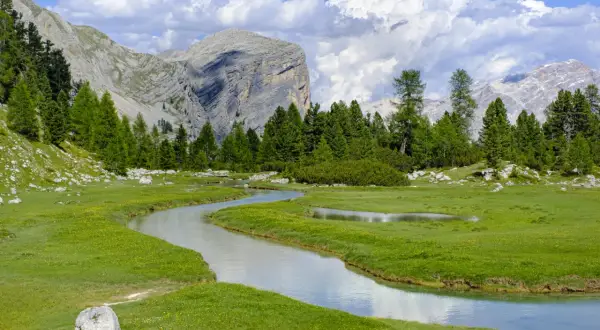 Un sereno sentiero di montagna serpeggia tra prati verdi e un ruscello cristallino, con imponenti vette sullo sfondo.