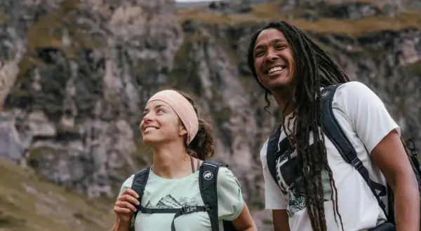 Coppia sorridente in montagna, con zaini e abbigliamento montagna estate perfetto per l'avventura.