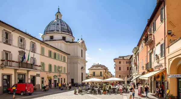 Piazza a Castel Gandolfo, dove si trova il Palazzo Apostolico. Edifici colorati, una chiesa con cupola e tavolini all'aperto animano la piazza.